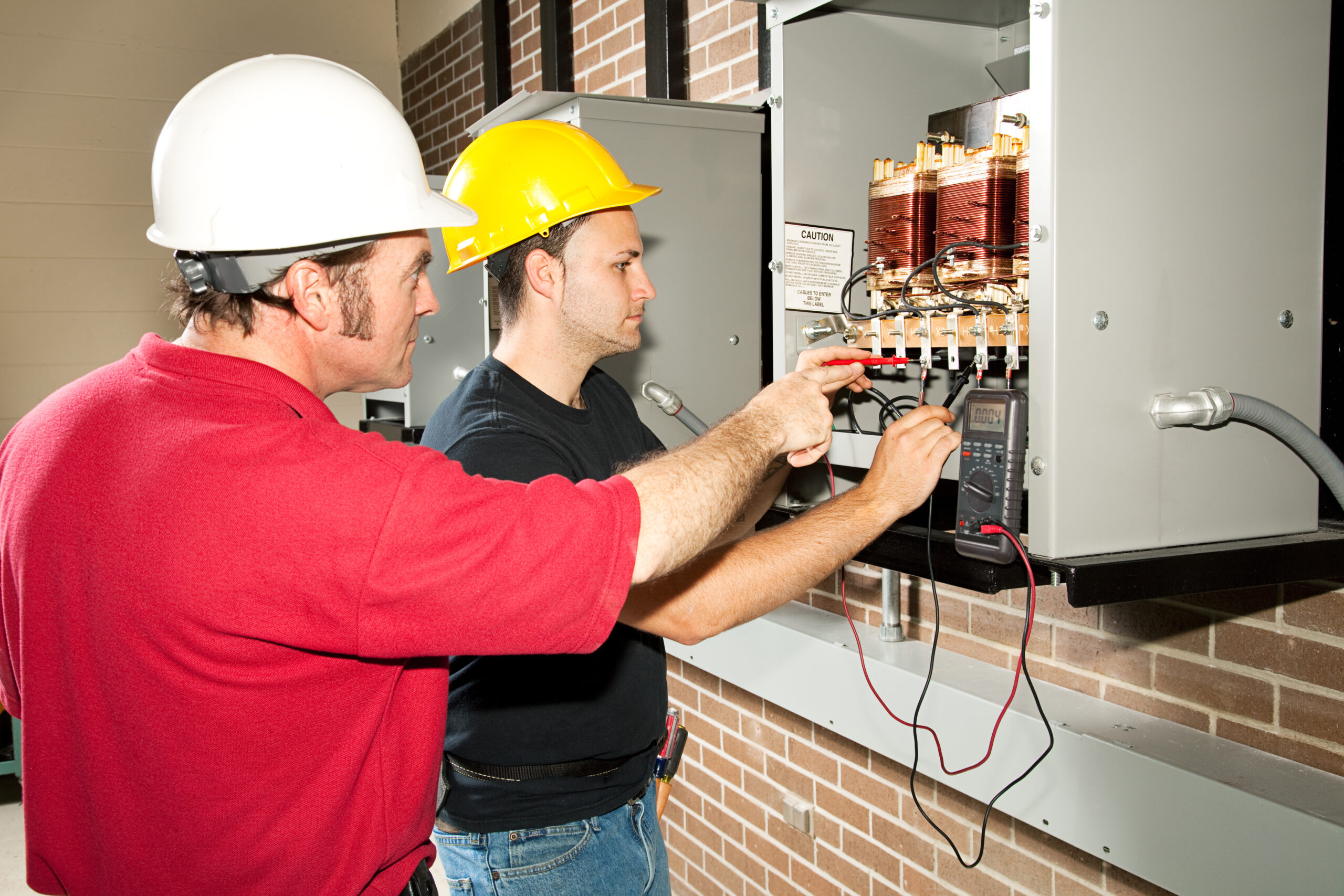 Journeyman electrician training an apprentice while testing a transformer with a digital multimeter.